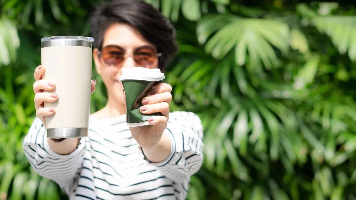 Frau hält einen wiederverwendbaren Coffee-to-go-Becher und einen zerdrückten Einweg-Becher in die Kamera